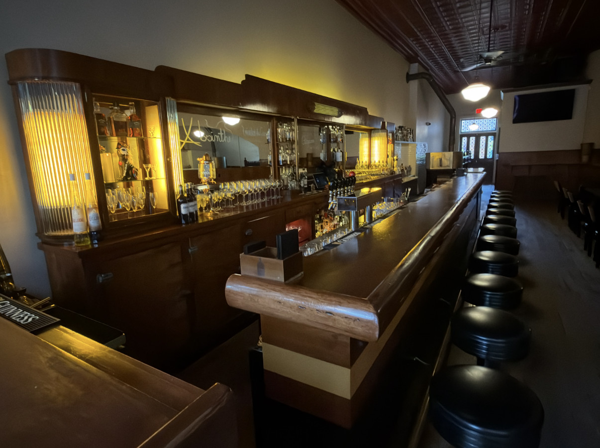 Long polished wooden bar lined with black leather stools, a glowing mirrored back bar and a pressed-tin ceiling with pendant lights