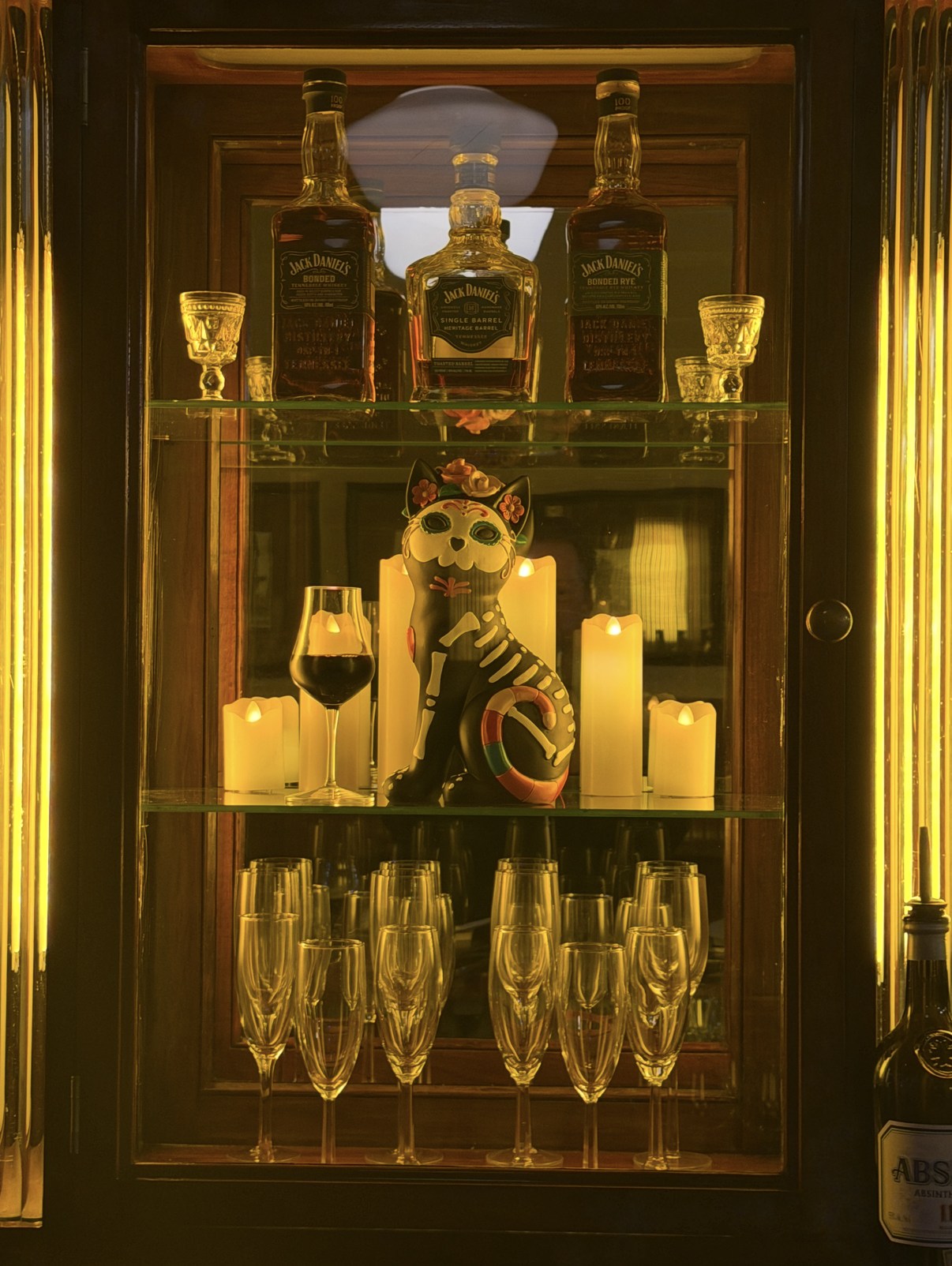 Lit glass cabinet with whiskey bottles, glowing candles, rows of champagne flutes and a sugar-skull cat figure