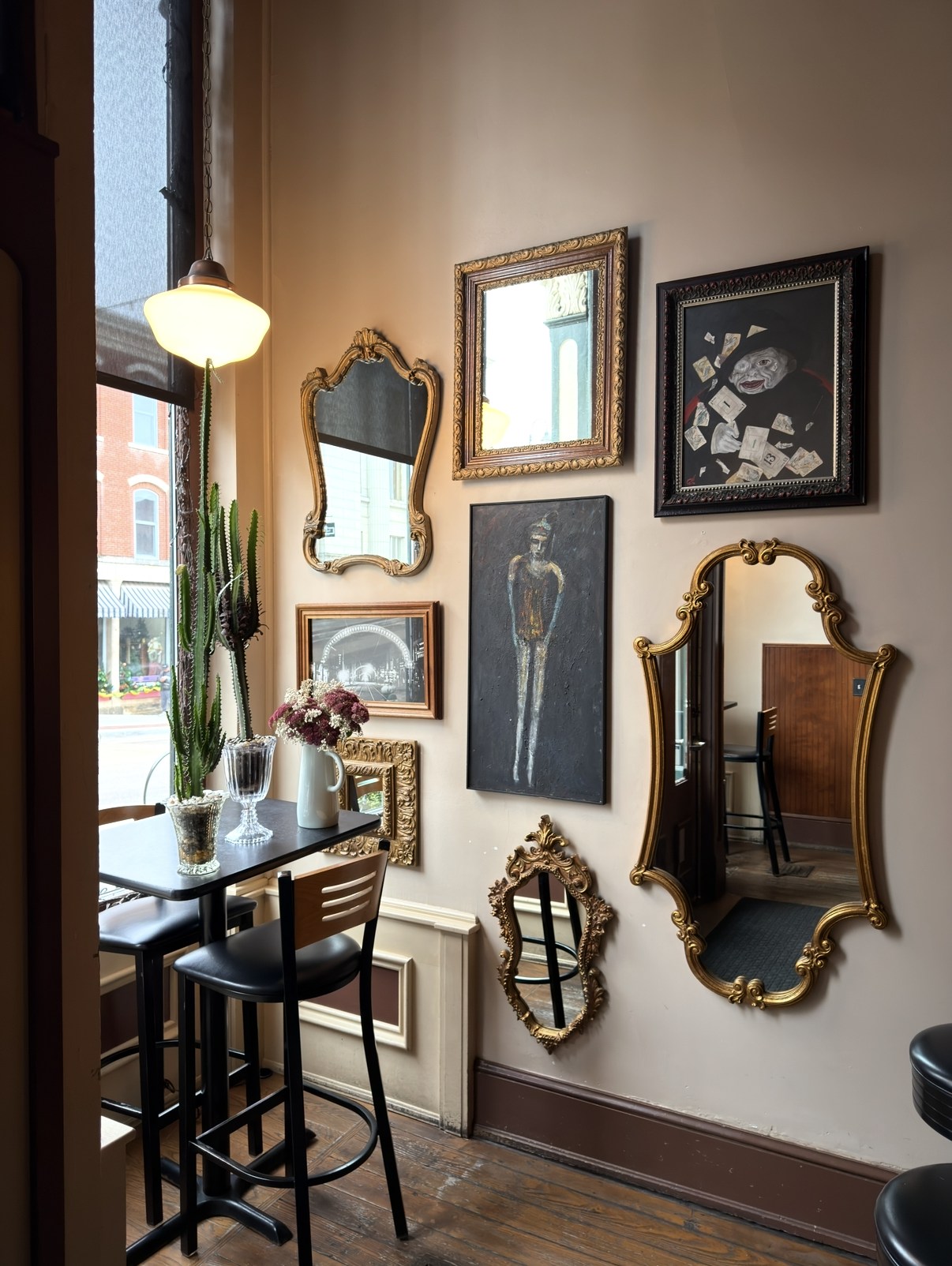 High-top table and bar stools by the front window beside a gallery wall of gilded mirrors and framed art