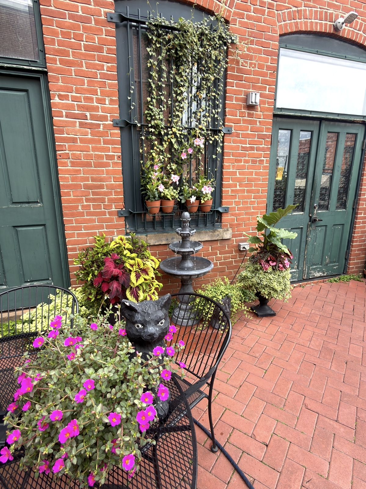Patio fountain beneath an ivy-covered barred window on the red brick wall, with a black cat statue and pink flowers on an iron table and herringbone brick pavers