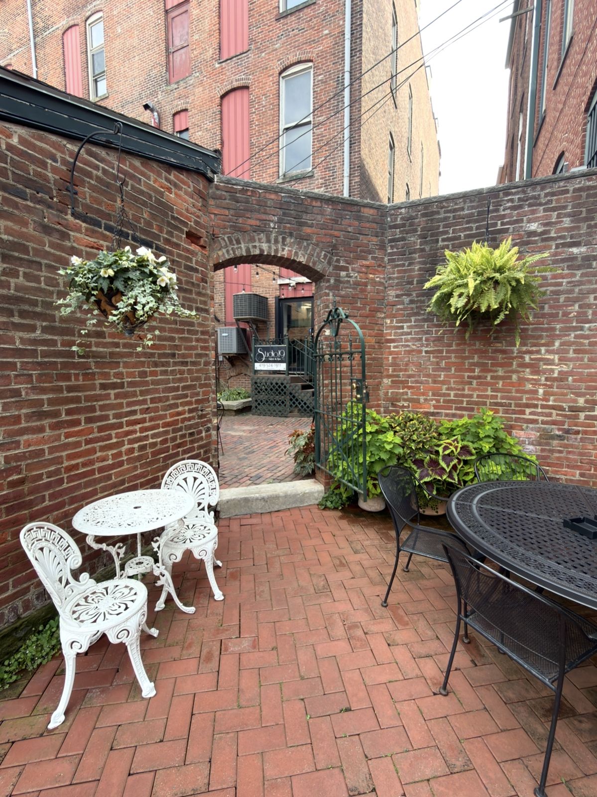 Walled brick courtyard with an arched gateway and iron gate, a white ornate bistro set and hanging plants