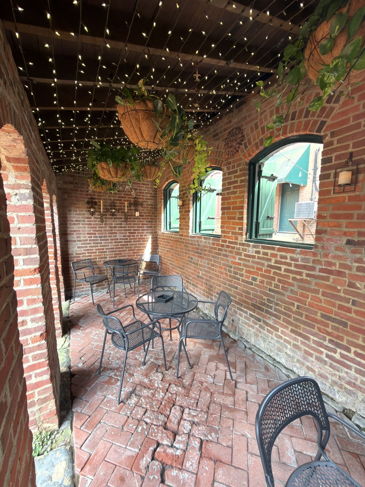 Covered brick arcade on the patio with string lights overhead, hanging baskets and black iron tables