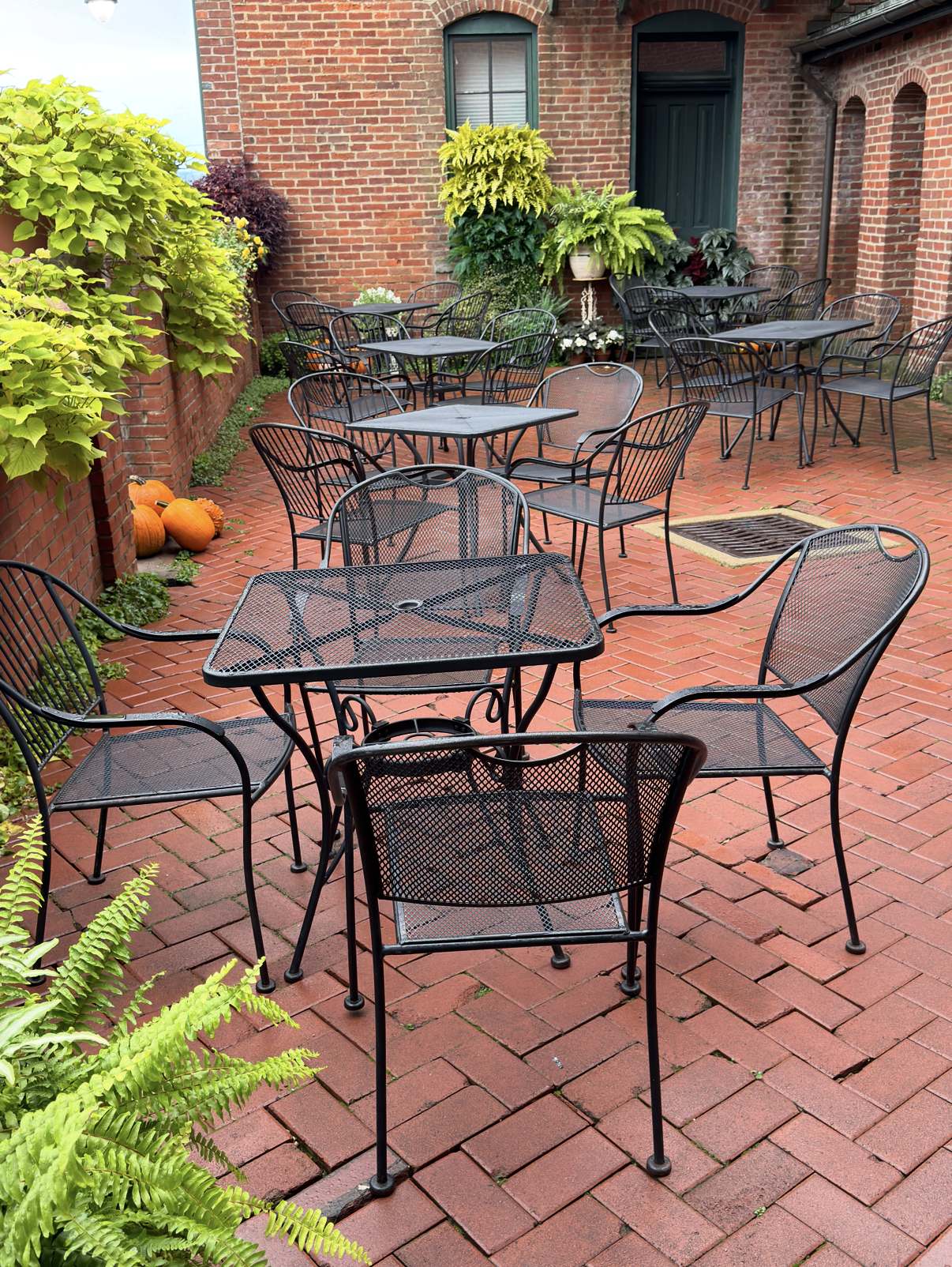 Black iron tables and chairs on herringbone brick pavers, surrounded by lush greenery, ferns and brick walls