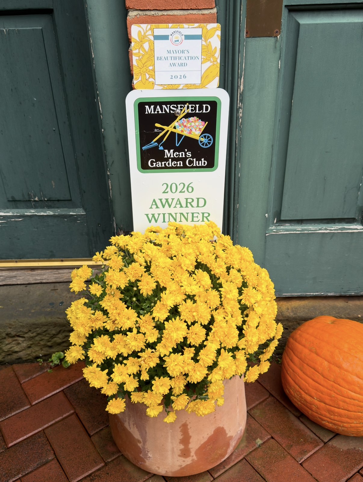 2026 Mayor's Beautification Award and Mansfield Men's Garden Club 2026 Award Winner signs beside a pot of yellow mums and a pumpkin at the bar's green door